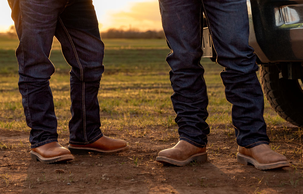 Close-up of two men wearing brown Homeland work boots and jeans, standing on dirt near a truck at sunset.